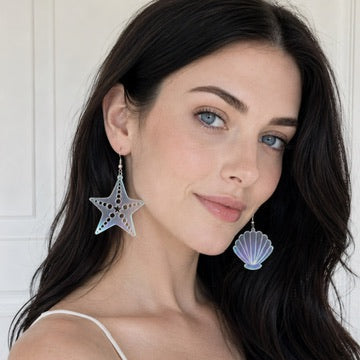 A woman with long dark hair wearing dangling earrings shaped like a starfish and a scallop shell, photographed against a light paneled wall.