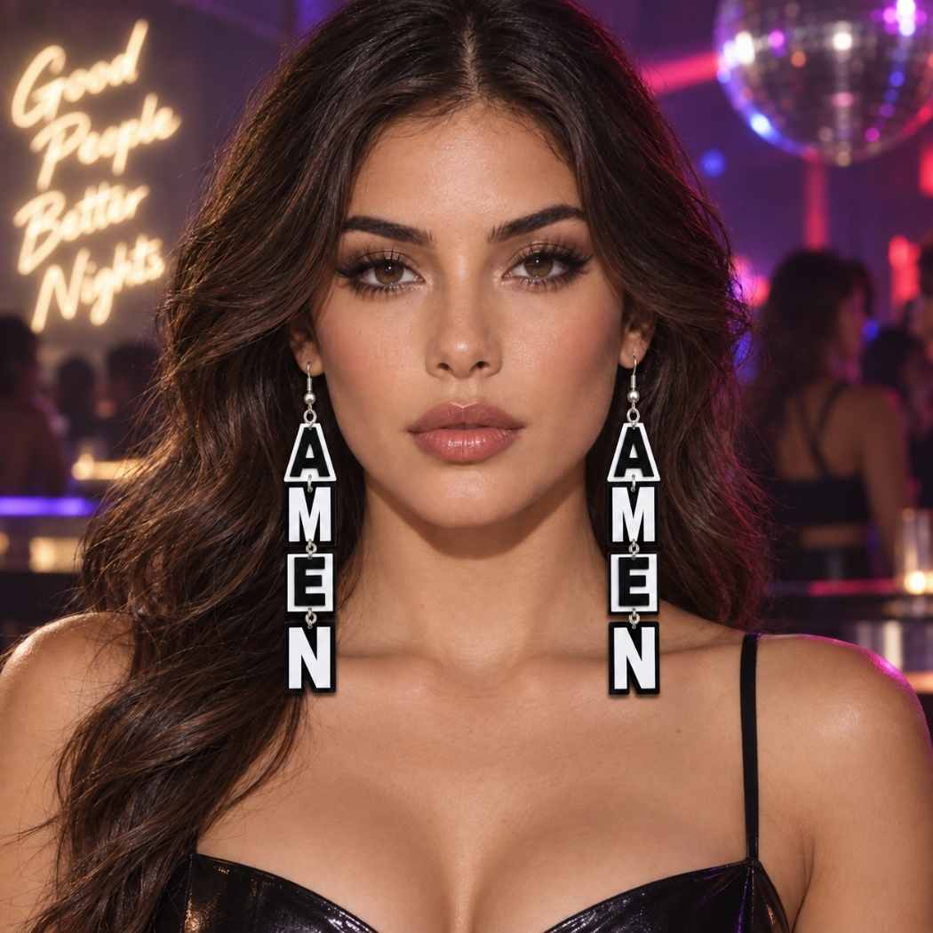 A woman in a nightclub setting wearing long dangling earrings made of stacked square tiles spelling 'AMEN' vertically. The background shows a disco ball and neon signs. Black and white AMEN statement acrylic drop earrings by SassyBirds