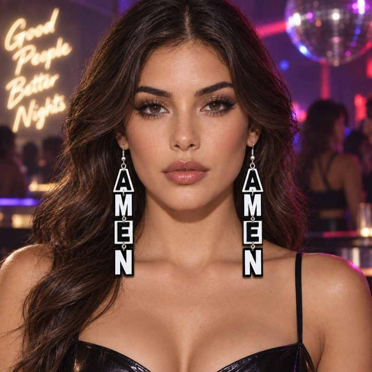 A woman in a nightclub setting wearing long dangling earrings made of stacked square tiles spelling 'AMEN' vertically. The background shows a disco ball and neon signs. Black and white AMEN statement acrylic drop earrings by SassyBirds