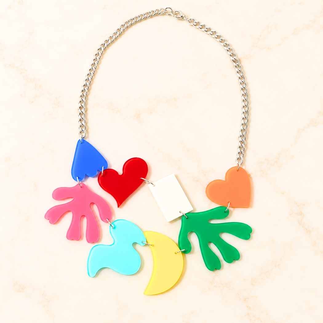 A colorful statement necklace featuring multiple acrylic charms in pink, blue, red, orange, green, yellow, and white, suspended from a silver chain on a light beige marble background.