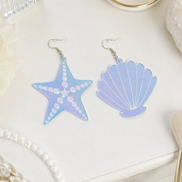 A pair of dangling earrings shaped like a starfish and a seashell, featuring a light blue and lavender gradient with white polka dots and lines, displayed on a white surface with decorative props.