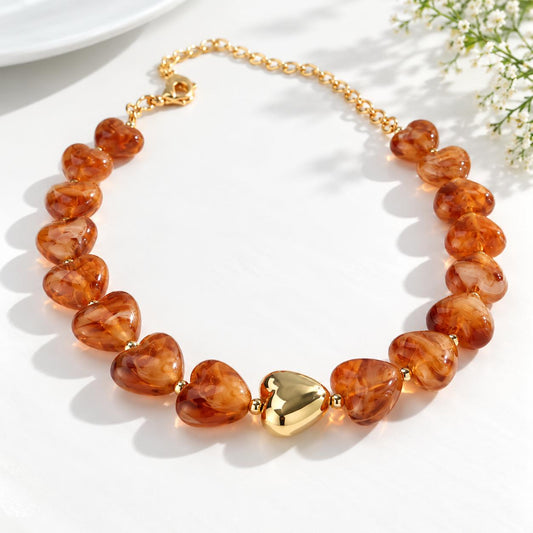 A heart-shaped beaded necklace made of translucent amber-orange stones with a polished gold heart in the center and a gold chain clasp, photographed on a white surface with soft shadows and small white flowers in the corner.