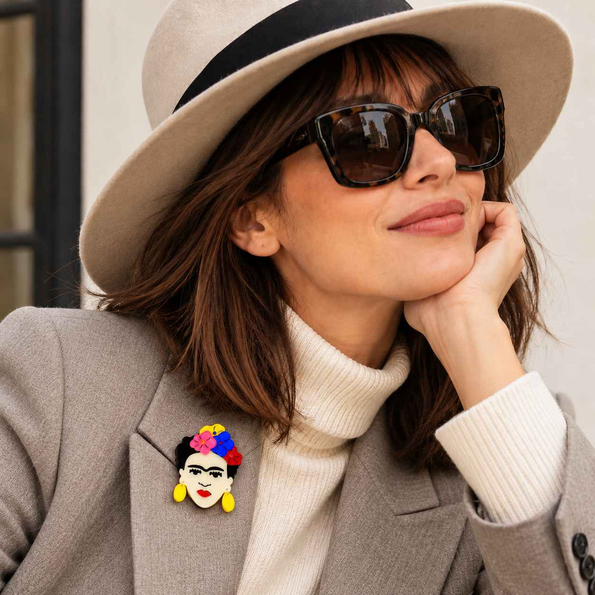 A woman wearing a beige wide-brim hat, dark sunglasses, a gray blazer, and a cream turtleneck, with a colorful Frida Kahlo-style face brooch pinned to her blazer.