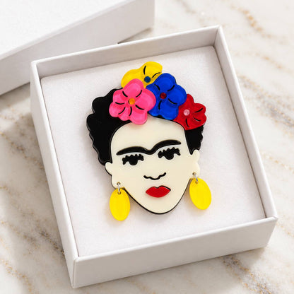 A stylized pin featuring a woman's face with black hair, a colorful flower crown in pink, blue, and red, red lips, and yellow earrings, displayed in a white gift box on a light marble surface.