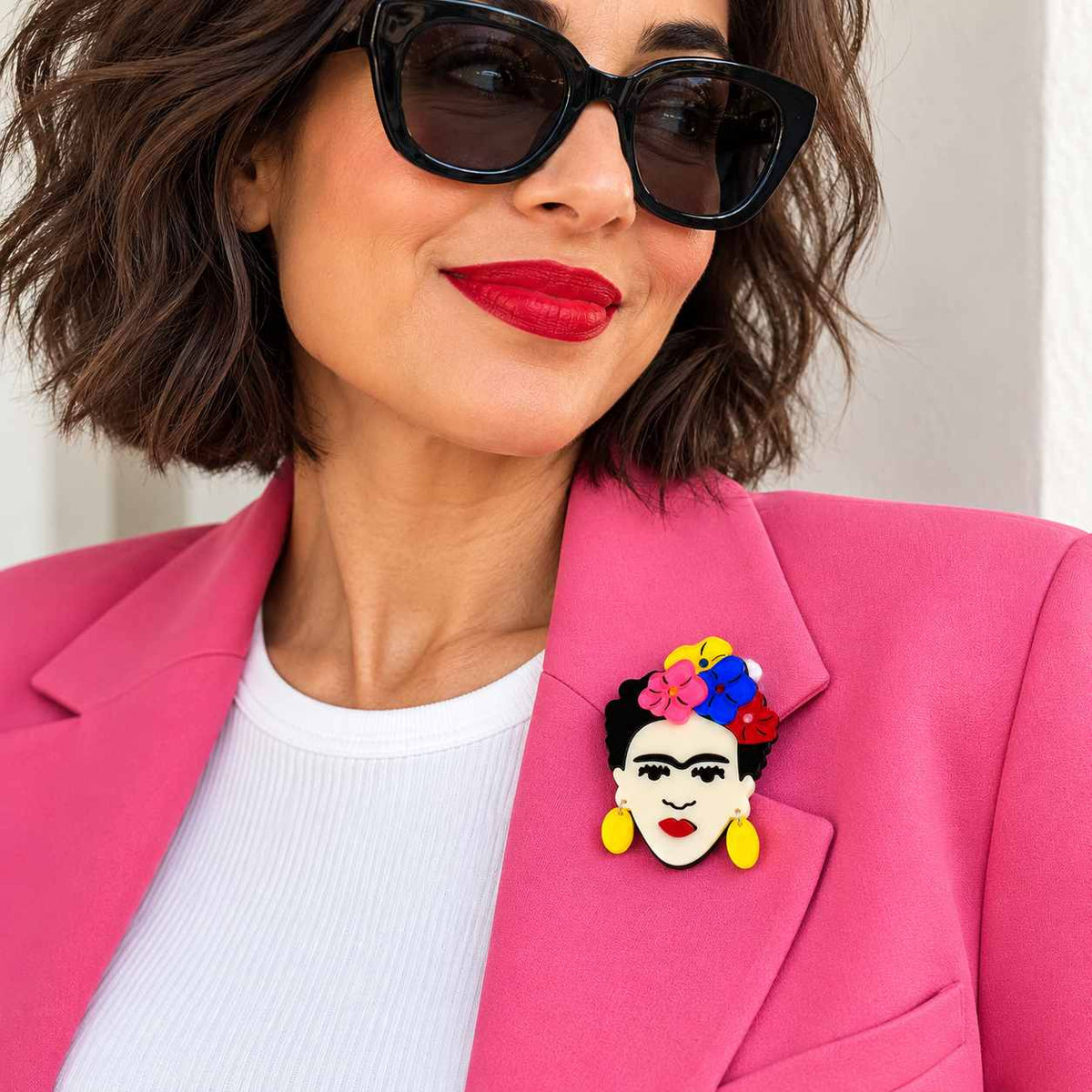 A close-up lifestyle shot of a woman wearing a colorful pin featuring Frida Kahlo's face and flowers on her collarbone, styled with a bright pink blazer and white top.