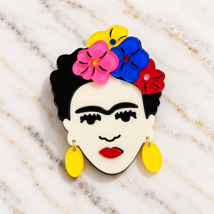 A colorful pin or charm shaped like a stylized Frida Kahlo face, with black hair, a white face, pink and blue flowers in the hair, a red flower, yellow earrings, and red lips, photographed on a light marble surface.
