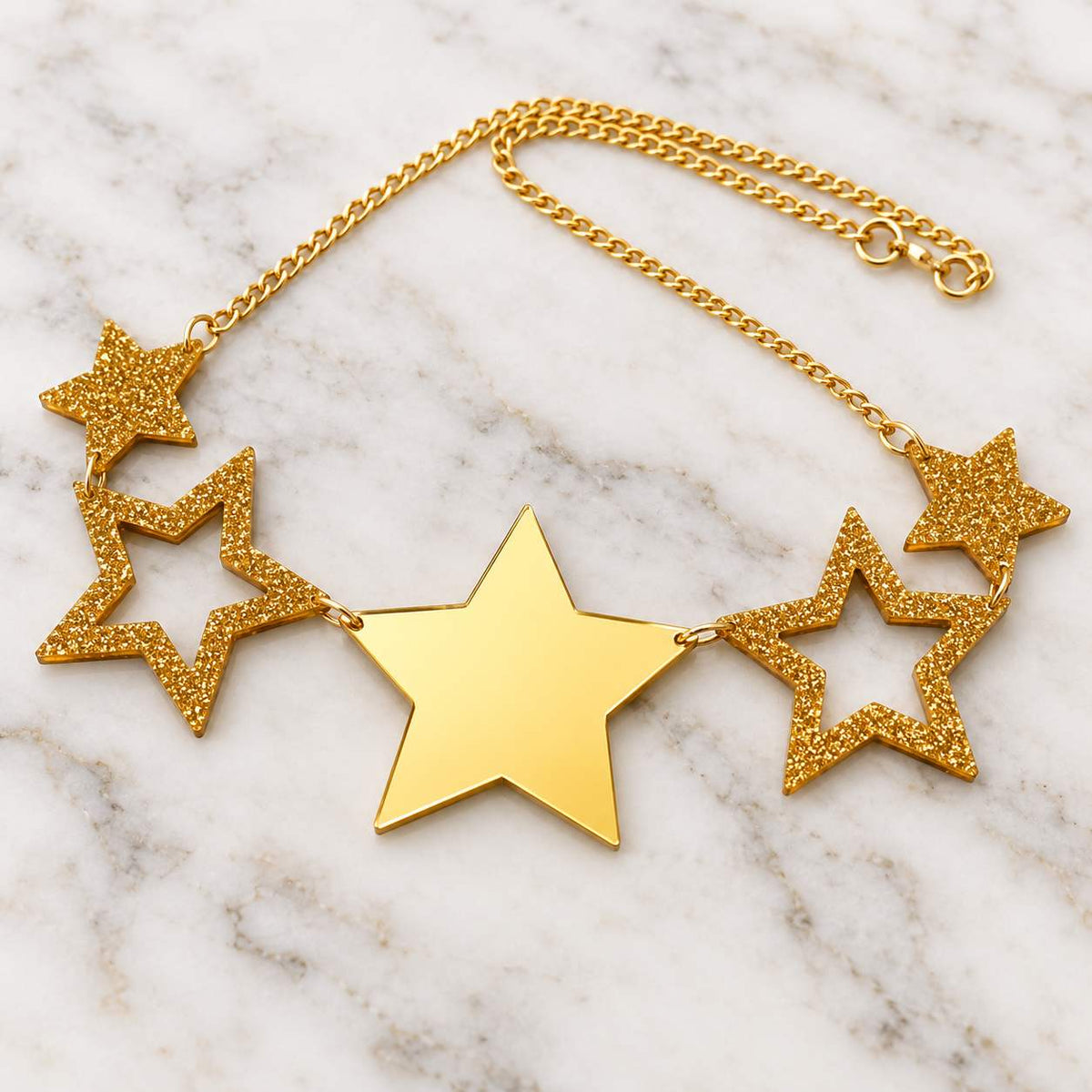 A gold star necklace featuring one large solid gold center star, two openwork glittery gold side stars, and two smaller glittery gold stars on the chain, displayed on a white marble surface.