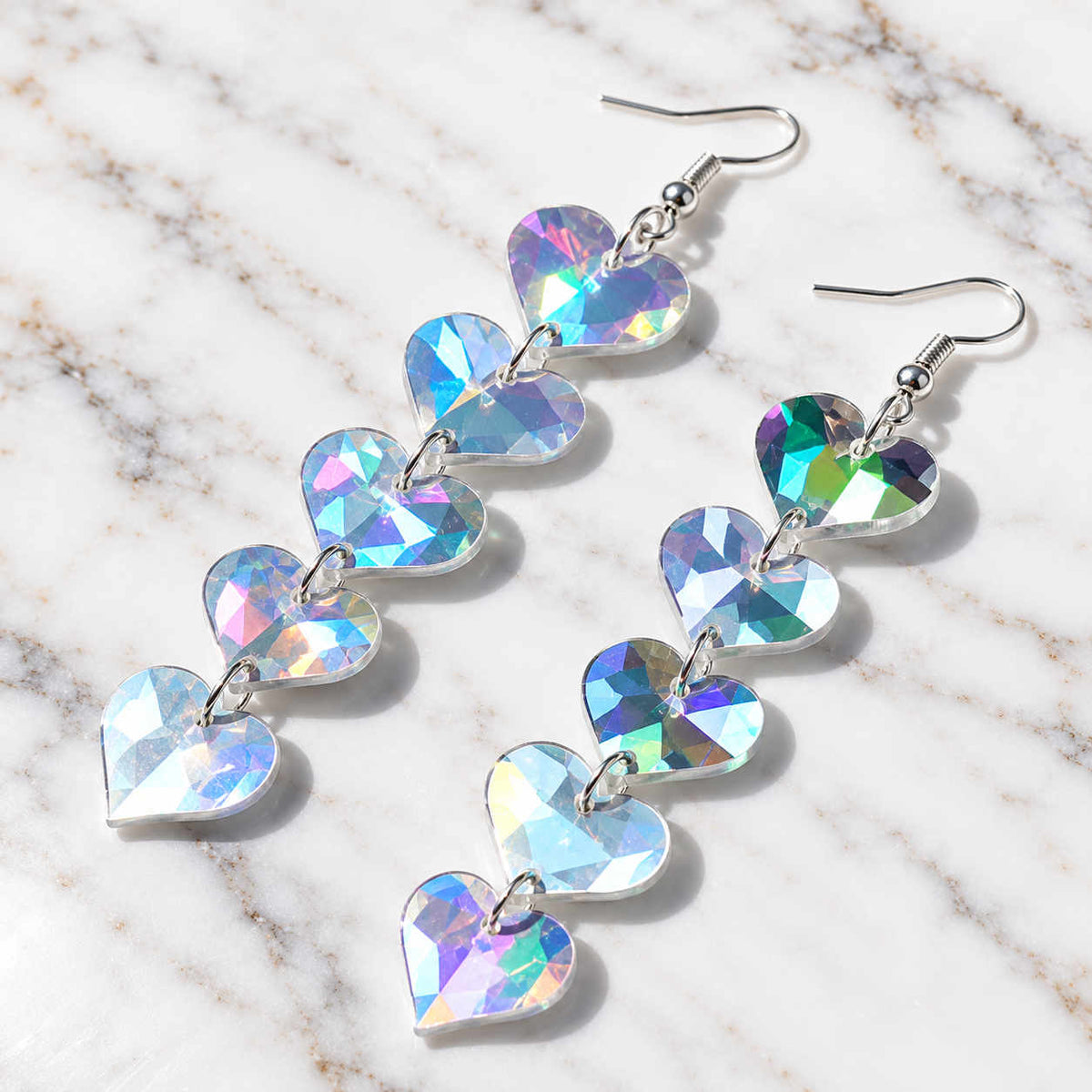 A pair of dangling heart-shaped crystal drop earrings with silver fishhook wires, displayed on a white marble surface. The crystals have an iridescent, holographic finish reflecting pastel pinks, blues, and greens.
