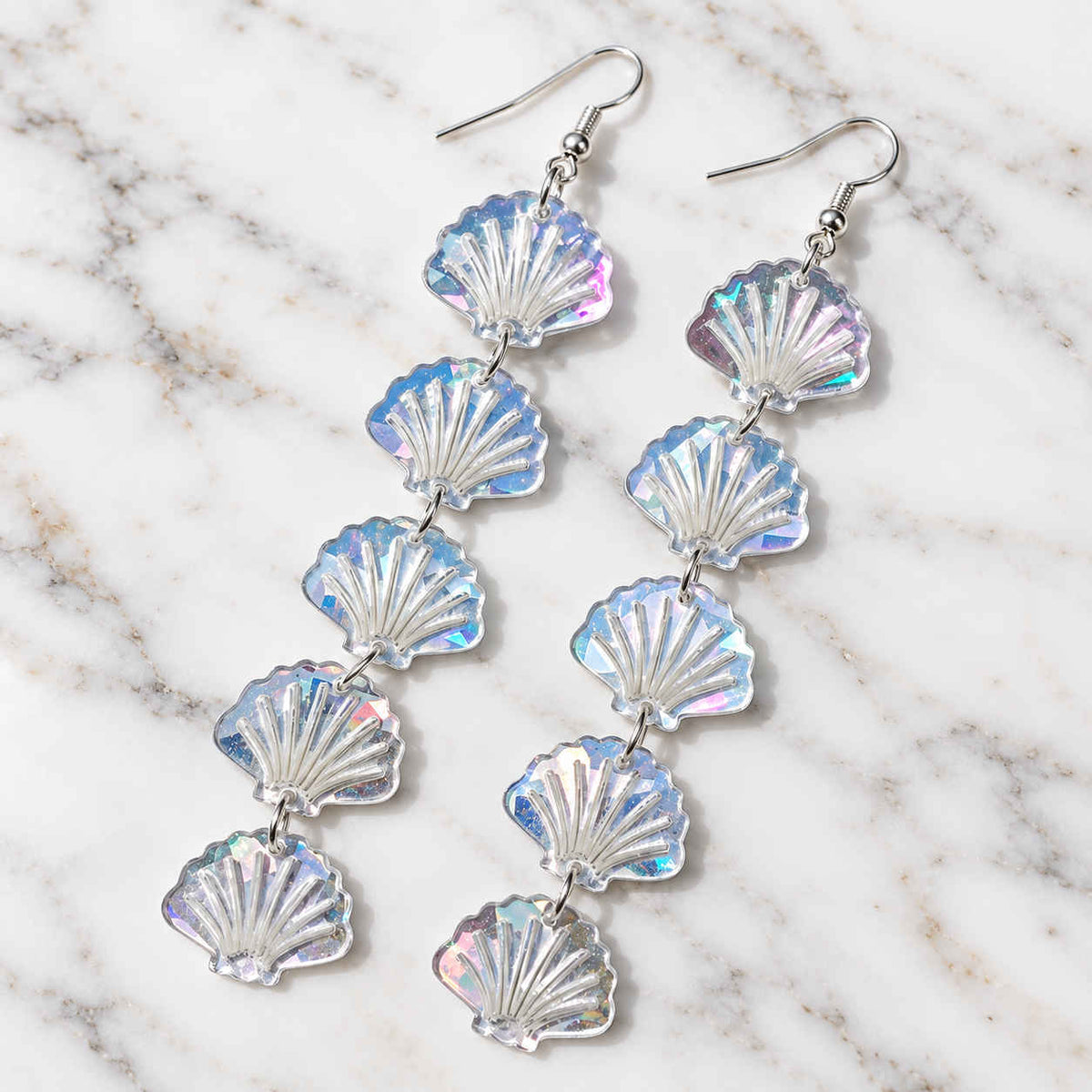 A pair of dangling shell-shaped earrings made of iridescent, translucent material with silver fishhook wires, displayed on a white and gray marble surface.