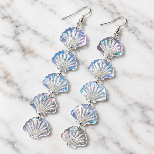A pair of dangling shell-shaped earrings made of iridescent, translucent material with silver fishhook wires, displayed on a white and gray marble surface.
